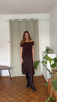 Abito marrone off-shoulder con un design arricciato, texture liscia e gonna a ginocchio, abbinato a stivali neri alti. Ambientazione naturale interna.