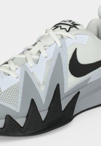 Άσπρο και γκρι αθλητικό παπούτσι με μαύρο ζιγκ-ζαγκ σχέδιο στην σόλα, μαύρο λογότυπο Nike Swoosh και λευκά κορδόνια σε απλό φόντο.