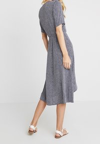 Marineblaues Kleid mit weißen Sprenkeln, kurzen Ärmeln, rundem Halsausschnitt und einem fließenden, knielangen Design. Kombiniert mit weißen Sandalen.