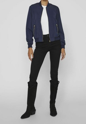 Vrouw met een marineblauwe bomberjack over een wit topje, zwarte strakke broek en zwarte kniehoge laarzen met hak, staande voor een effen achtergrond.