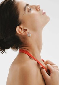 Pendientes de aro plateados con un diseño retorcido, adornados con pequeños embellecimientos, llevados en la oreja izquierda de una persona con un fondo neutro.
