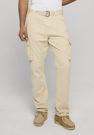 Uomo con pantaloni cargo beige con tasche laterali, cintura beige e scarpe casual marroni chiaro, in piedi davanti a uno sfondo grigio uniforme.