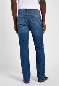 Dark blue denim jeans with a straight cut, visible stitching, two back pockets, and a brown leather logo patch at the waistband.