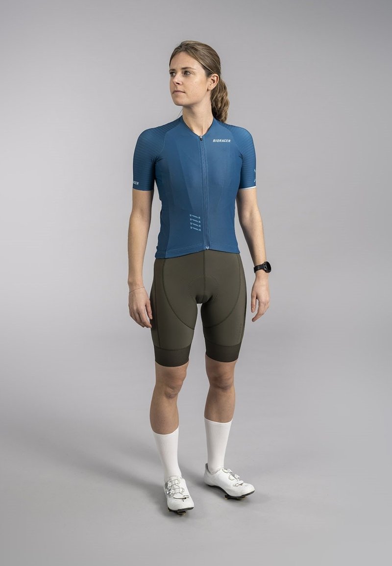 Blue cycling jersey with short sleeves, zip front, and textured design. Olive green padded shorts; white socks; white cycling shoes.