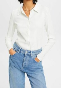 Vit skjorta med lång ärm och knappar, med vertikal ribbad textur, tillsammans med högmidjade blå denimjeans. Krage och knapprar synliga.