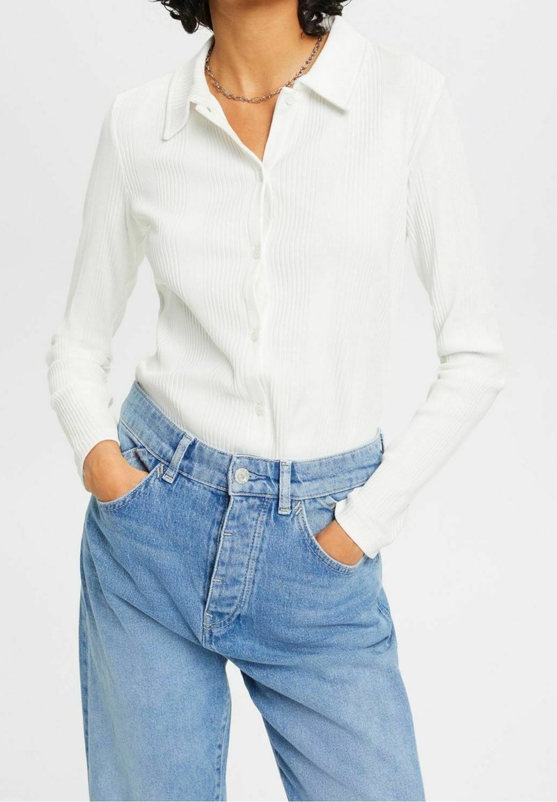 Vit skjorta med lång ärm och knappar, med vertikal ribbad textur, tillsammans med högmidjade blå denimjeans. Krage och knapprar synliga.