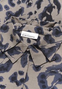 Cămașă cu guler dintr-un material bej estompat, cu un print animalier albastru și negru. Eticheta indică "KAREN BY SIMONSEN." Detaliu mic cu nasture vizibil.