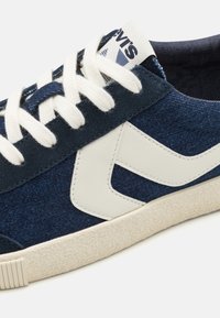 Kék denim sportcipő fehér bőr kiegészítővel. Texturált gumi talppal és krémszínű fűzőkkel rendelkezik. A logó a nyelven látható.