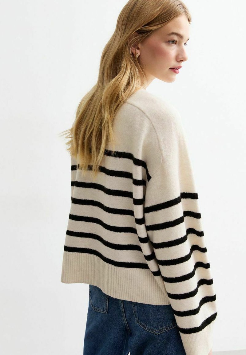 Maglione beige con strisce orizzontali nere, colletto a giro, e orlo a coste. Indossato con jeans in denim blu, che mostrano una vestibilità rilassata e oversize.