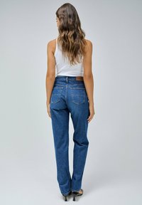 Jeans de mezclilla azul de cintura alta con corte recto, bolsillos traseros visibles y parche de cuero en la cinturilla, combinados con una camiseta sin mangas blanca.