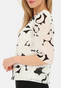 Biała bluzka z czarnymi abstrakcyjnymi wzorami, krótkie rękawy i luźny krój. Lekka tkanina o gładkiej fakturze.