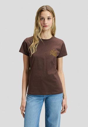 Frau steht, trägt ein braunes Lee Jeans 1889 T-Shirt und blaue Jeans, blickt nach vorne vor einem einfarbigen hellgrauen Hintergrund.