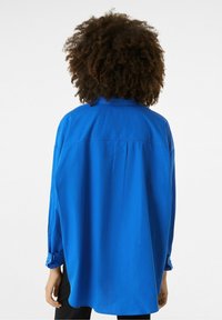Camisa azul de botones con mangas largas, dobladillo curvado y una abertura en la parte trasera para un mejor ajuste. Fabricada con un tejido suave y ligero.