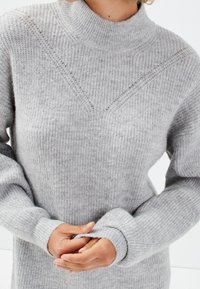 Pull gris en maille avec un col montant, une texture côtelée et un motif en chevron sur la poitrine. Présente des manches larges avec des poignets retroussés.