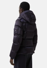Námořnická puffer bunda s prošívaným designem, velkou kapucí a zipovou kapsou na levém rukávu. Vyrobena z lesklého, odolného materiálu.