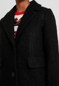 Manteau en laine noire avec une finition texturée, col cranté, poches avant et une fermeture à un bouton, superposé sur un pull rayé coloré.