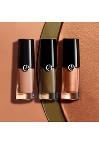 Tre flaconi di smalto per unghie Giorgio Armani in tonalità nude, verde oliva e rame metallizzato su superfici di colore abbinato.