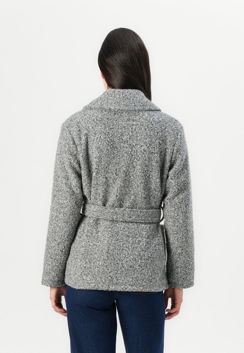 Veste enveloppante grise et texturée avec un large col et un détail de ceinture. Le tissu est doux, avec un aspect duveteux, complété par des manches longues.