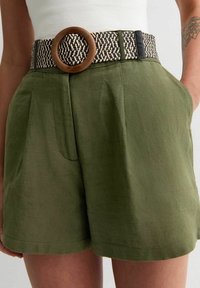 Olivgrüne Shorts mit Falten, versehen mit einem breiten gewebten Gürtel mit einer runden Holzschnalle und schwarzen Akzenten. Texturdetails des Stoffs sind sichtbar.