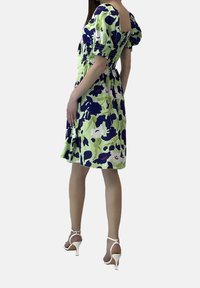 En grön blommig klänning med puffärmar har en fyrkantig rygg och smockad midja, i kombination med vita remsandaler med hög klack.