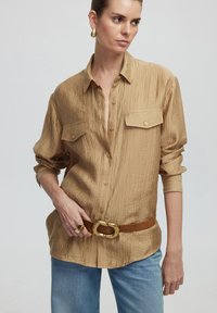 Beige zerknittertes Hemd mit Knopfverschluss, zwei Brusttaschen, hochgekrempelten Ärmeln, kombiniert mit einem braunen Gürtel mit einer goldfarbenen Schnalle.