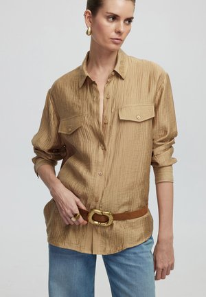 Beige crinkled shirt with button-down front, two chest pockets, rolled sleeves, paired with a brown belt featuring a gold-tone buckle.