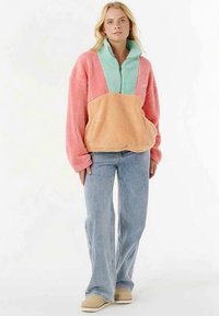 Fleece pullover in pink, mint, and peach. Features a quarter-zip collar and large front pocket. Paired with light blue jeans and beige shoes.