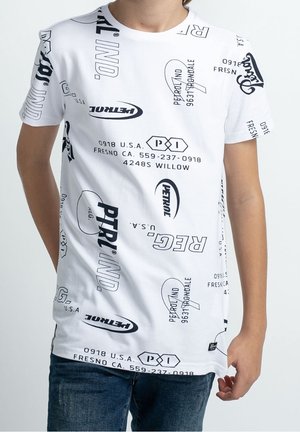 Camiseta blanca de manga corta con textos y logotipos variados en negro, llevada por una persona con una mano relajada a su lado.