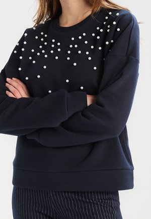 Femme portant un pull bleu marine décoré de petites perles blanches dispersées sur la poitrine et les épaules, les bras croisés, associé à un pantalon à fines rayures.