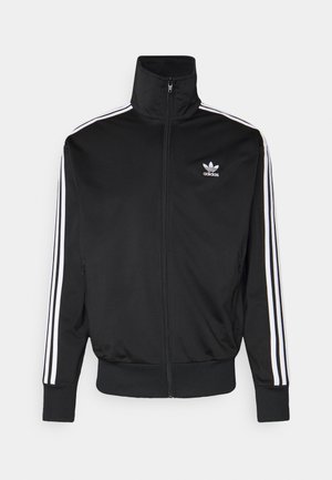 Persoon draagt een zwart Adidas-jasje, wit T-shirt, blauwe spijkerbroek, rode bandana en zwart-witte sneakers, staand op een straat.