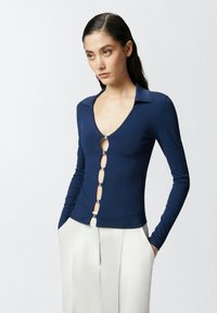 Mornarica dugi rukav majica s dubokim V-izrezom i srebrnim detaljima. Ima zatvaranje s lancem sprijeda. Glatka, rastezljiva tkanina.