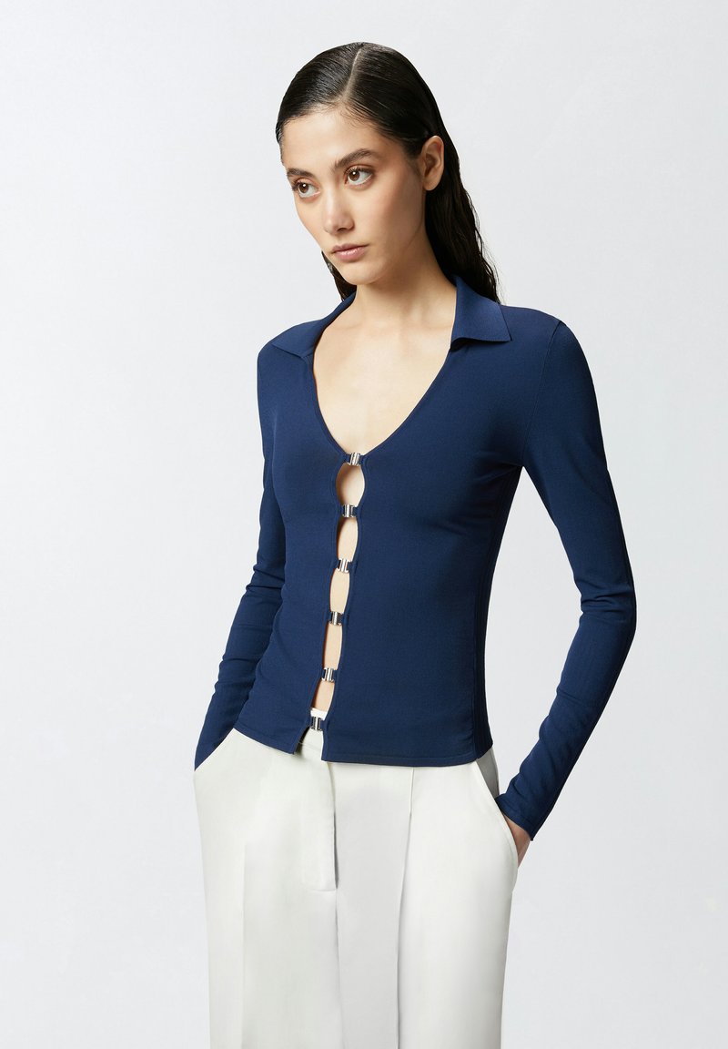 Mornarica dugi rukav majica s dubokim V-izrezom i srebrnim detaljima. Ima zatvaranje s lancem sprijeda. Glatka, rastezljiva tkanina.