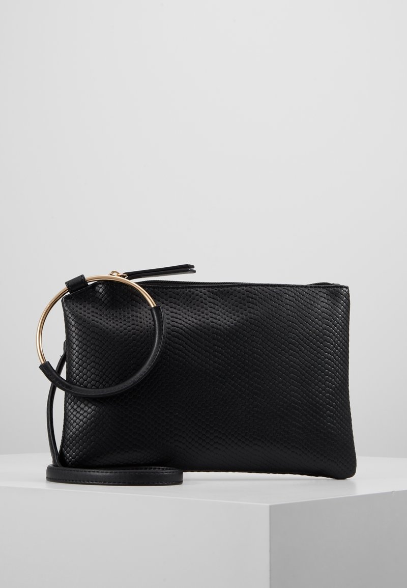 Pochette noire texturée avec une poignée circulaire dorée et une sangle de poignet détachable sur une surface blanche.