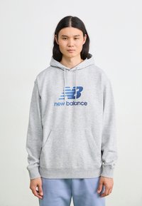 Siv hoodie z veliko sprednjo žepe, kapuco na vrvico in modrim logotipom 'New Balance'. Mehka tkanina s sproščenim krojem in rebrastimi manšetami.