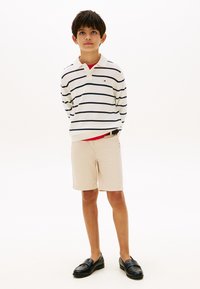 Αγόρι που στέκεται με τα χέρια πίσω από την πλάτη, φορώντας λευκό μακρυμάνικο πόλο με μαύρες ρίγες, μπεζ σορτς και μαύρα παπούτσια τύπου slip-on.