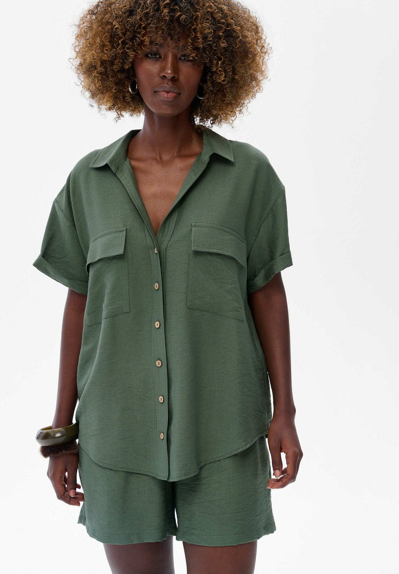 Vrouw met krullend haar, gekleed in een losse groene knopenoverhemd en bijpassende shorts, geaccessoriseerd met armbanden om haar linkerpols, staande tegen een witte achtergrond.