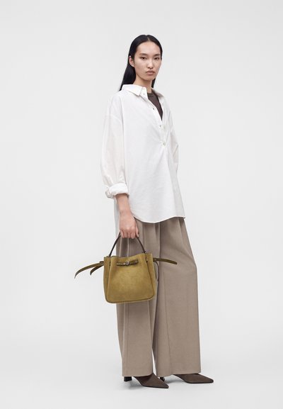 Femme portant une chemise blanche oversize, un pantalon large beige et des chaussures marron pointues tenant un sac à main en suède beige.