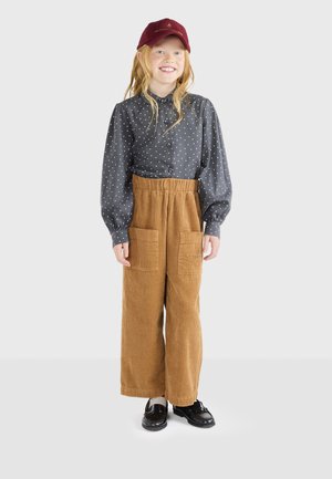 Camisa gris con lunares, botón frontal, pantalones de pana marrón de pierna ancha con bolsillos y gorra burdeos. Unos zapatos negros brillantes completan el conjunto.