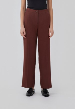 Osoba nosząca brązowe spodnie z wysokim stanem i szerokimi nogawkami, w połączeniu z czarnymi mokasynami i czarnym bezrękawnikiem, stojąca na jednolitym tle.