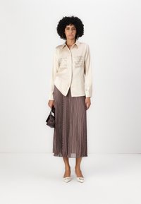 Beige szatén ing, két díszített zsebbel, amelyet egy lengő barna szoknyával párosítanak, amely bögrék mintájú. A modell egy kis sötét táskát tart.
