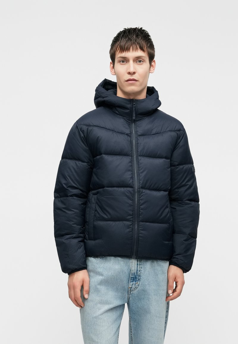 Chaqueta abrigada de color azul marino con capucha, que presenta secciones acolchadas y un cierre frontal. Combinada con unos vaqueros de denim azul claro.