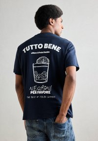 Marineblå T-skjorte med et hvitt grafisk trykk av et cocktailglass med is og en appelsinskive. Teksten sier "TUTTO BENE" og "NEGRONI PER FAVORE."