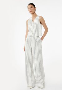 Frau in einem weißen ärmellosen, geknöpften Vest und weiten Hosen mit dünnen schwarzen vertikalen Streifen, steht vor weißem Hintergrund.