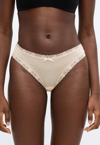 Γυναίκα που φοράει μπεζ εσώρουχο με δαντέλα και μικρό φιόγκο στο μπροστινό μέρος, και μαύρο κοντό τοπ, όρθια με χαλαρωμένα τα χέρια.