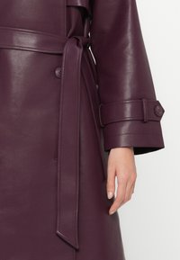 Long, deep purple leather coat with a belted waist, button accents on the cuffs, and a smooth, glossy texture.