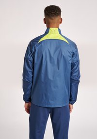 Marinblå regnjacka med hög krage, gula accenter på axlarna och strukturerade paneler. Lätt, vattenavvisande tyg med elastiska ärmslut.