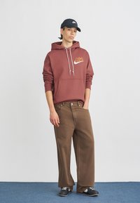 Bordový mikina s bielym logom Nike a šnúrkami, spárovaná s voľnými hnedými nohavicami a čiernymi topánkami, na jednofarebnom pozadí.