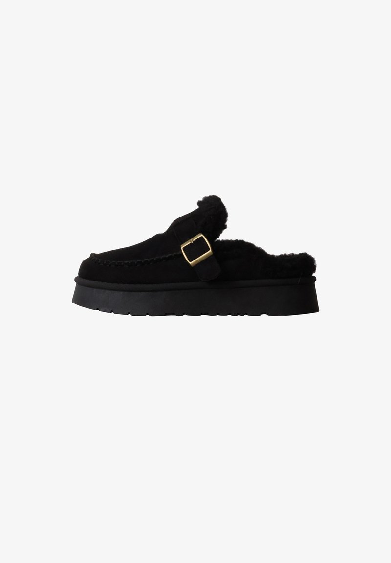 Mocassin noir à enfiler avec semelle épaisse, bride avec boucle dorée et doublure en fourrure douce visible autour de l'ouverture.