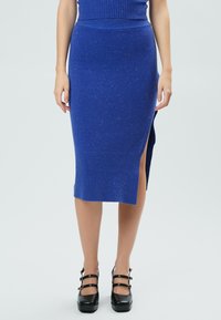 Blaue Strickrock mit einem gerippten Taillenbund, ausgestattet mit einem seitlichen Schlitz. Der Stoff hat dezente Farbnuancen und eine glatte Textur. Kombiniert mit schwarzen Plateauschuhen.