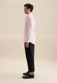 Lichtroze blouse met lange mouwen en een borstzak, gecombineerd met donkerblauwe broek en zwarte schoenen, met een klassieke kraag en knoopjesmanchetten.